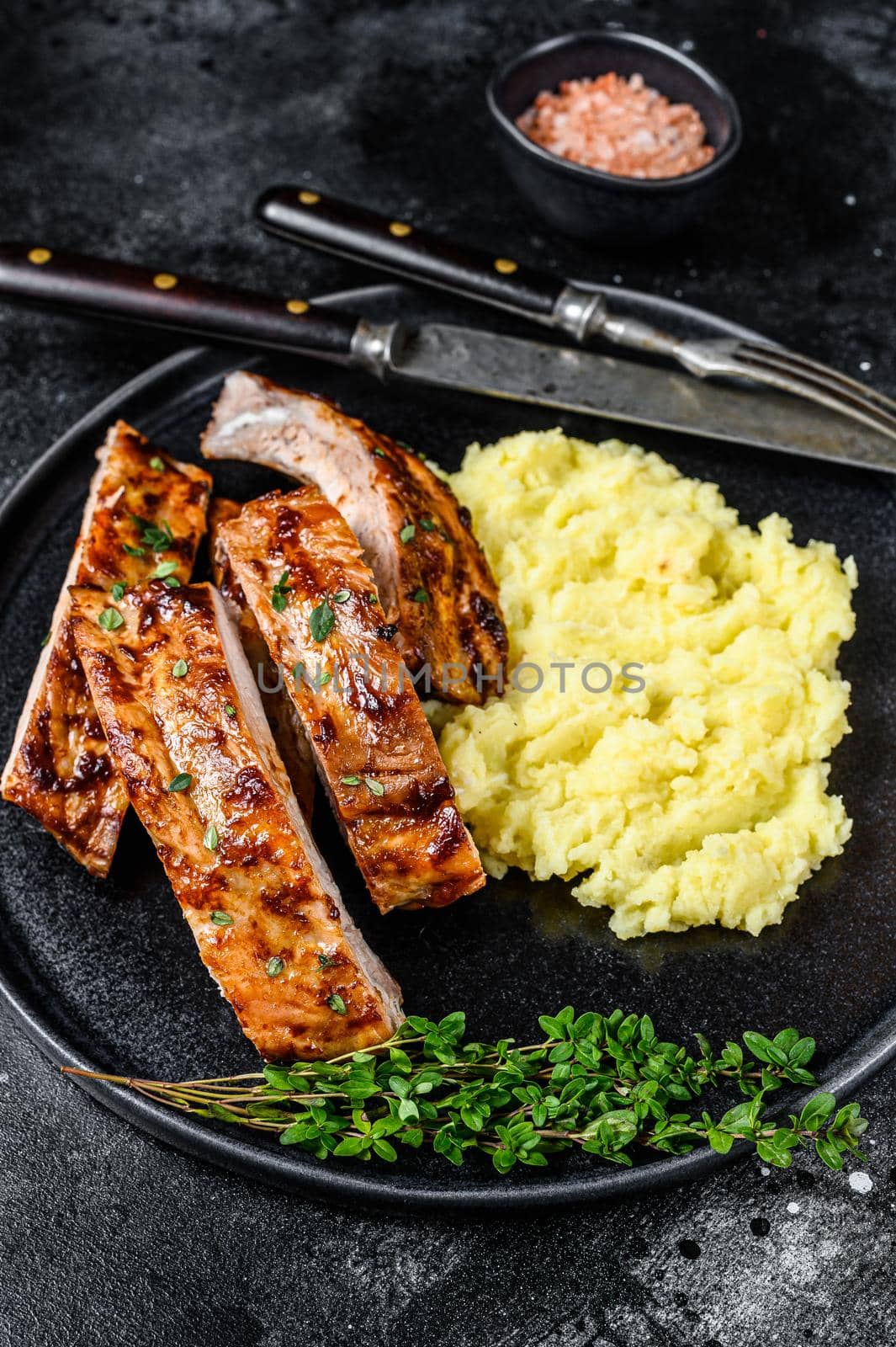 Sliced BBQ grilled pork rack spare ribs on a plate with mash potato. Black background. Top view by Composter