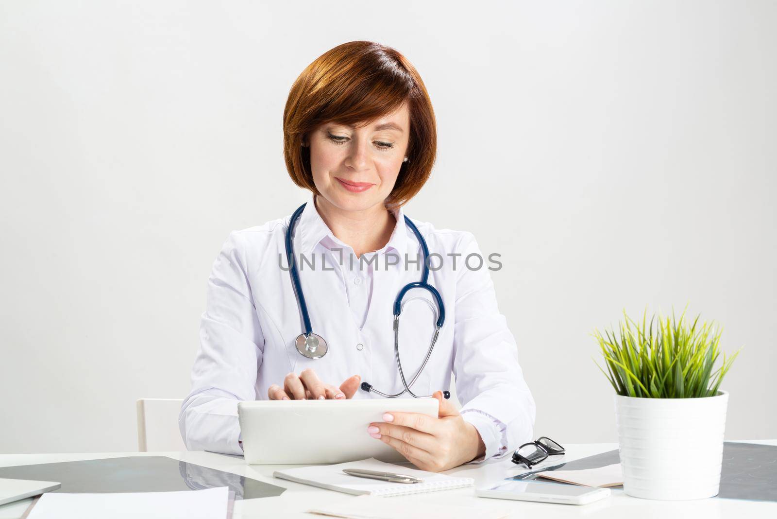 Beautiful doctor using tablet computer in office. Family physician in white coat with stethoscope sitting at desk. Professional medical diagnosis and treatment in clinic. Healthcare and medicine.