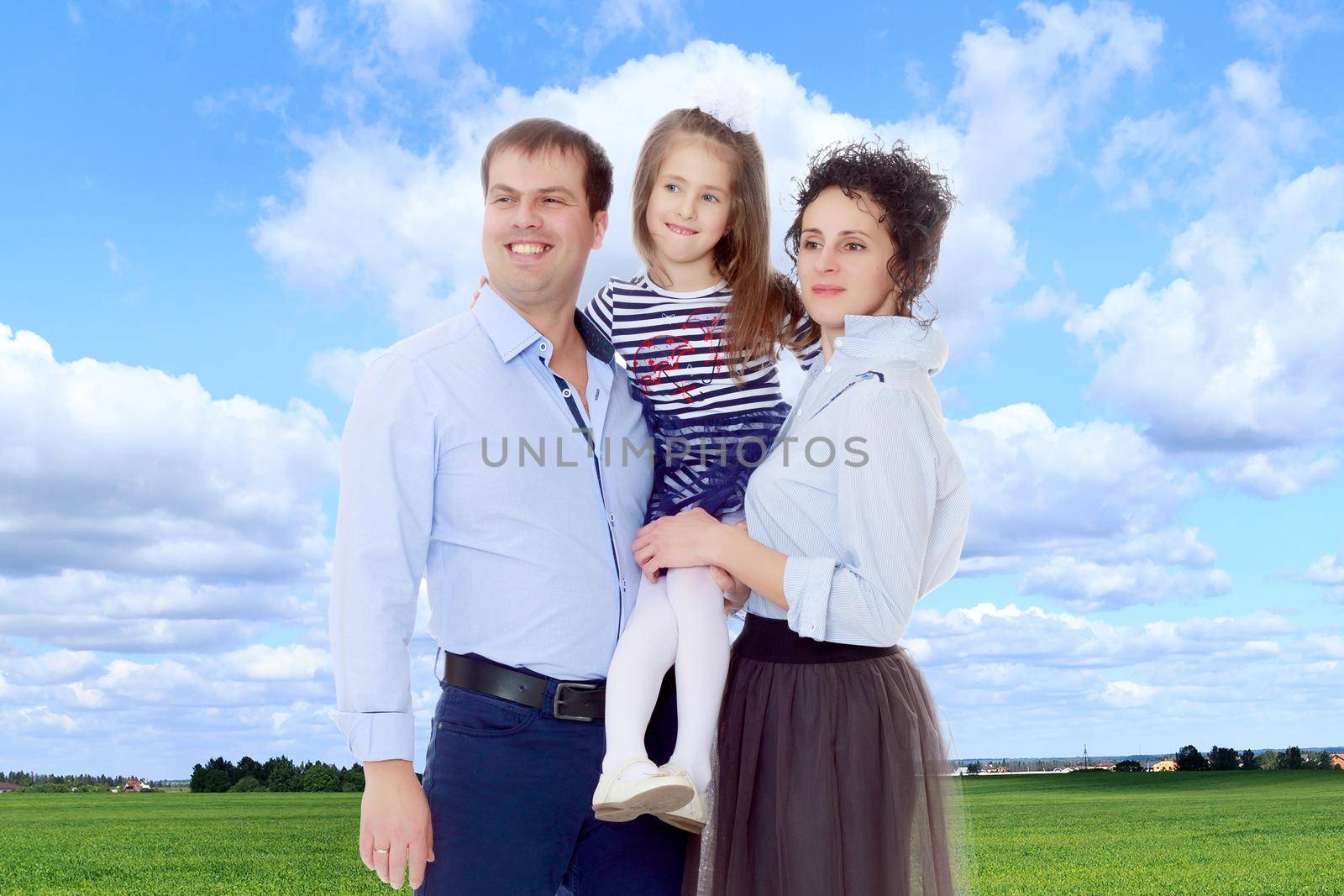 Happy young family, mom dad and little daughter.Parents keep the girl in her arms , and she hugs their neck.On the background of green grass and blue sky with clouds.