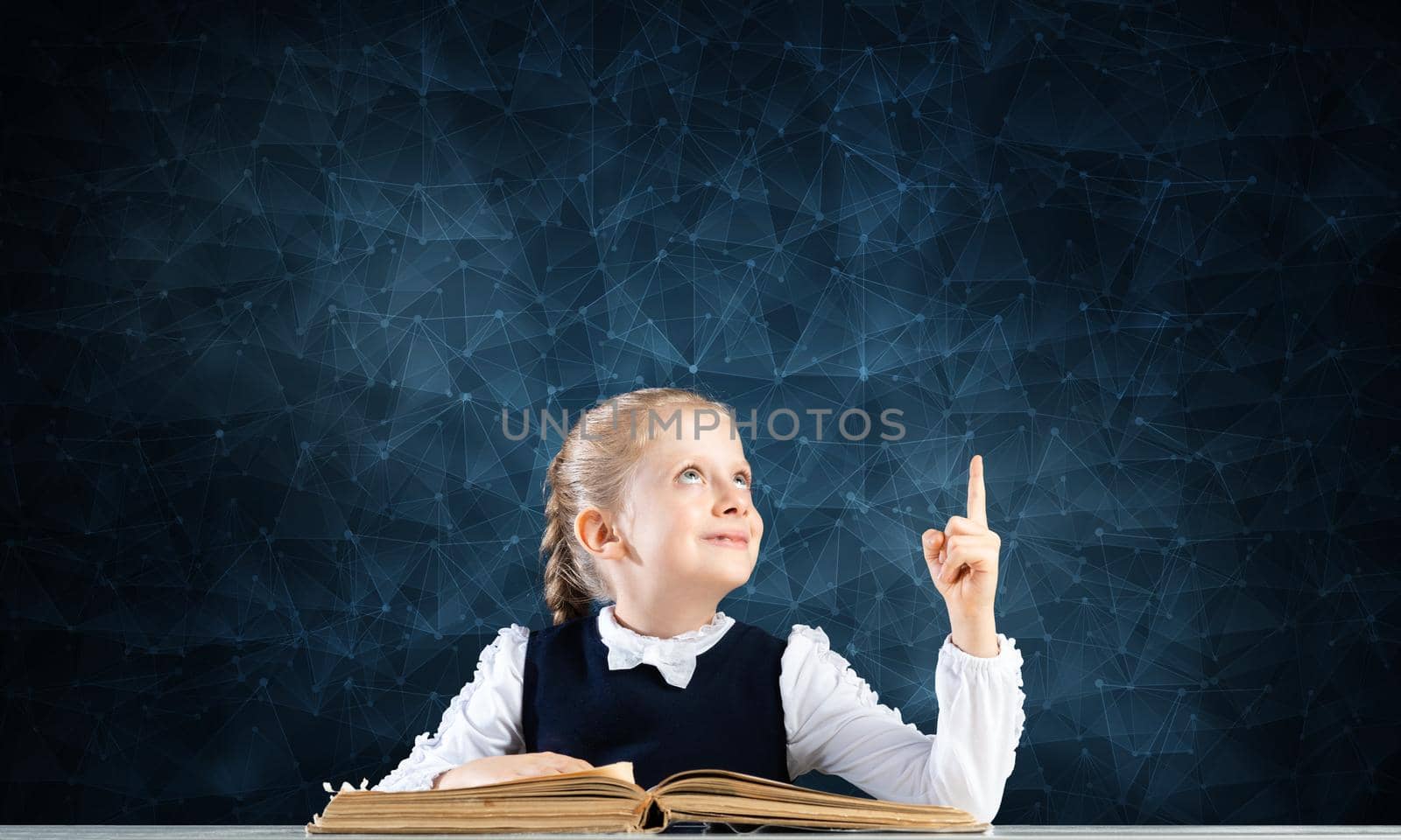 Smiling little girl sitting at desk with open book. Research and education in school. Happy schoolgirl in school wear finger pointing up. Clever girl reading book on deep blue plexus background