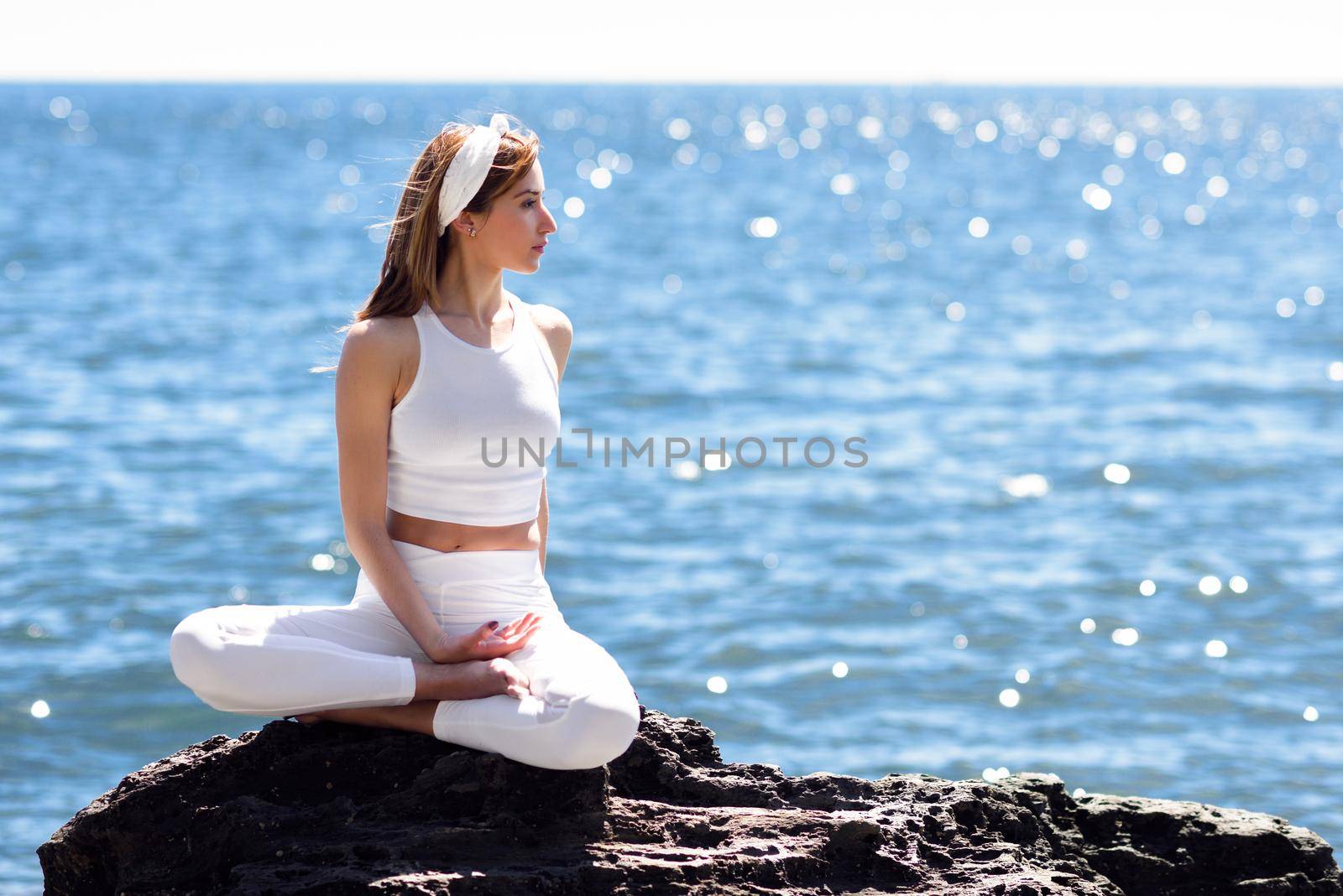 Young woman doing yoga in the beach. Female wearing white sport clothes in lotus figure with defocused sea background.