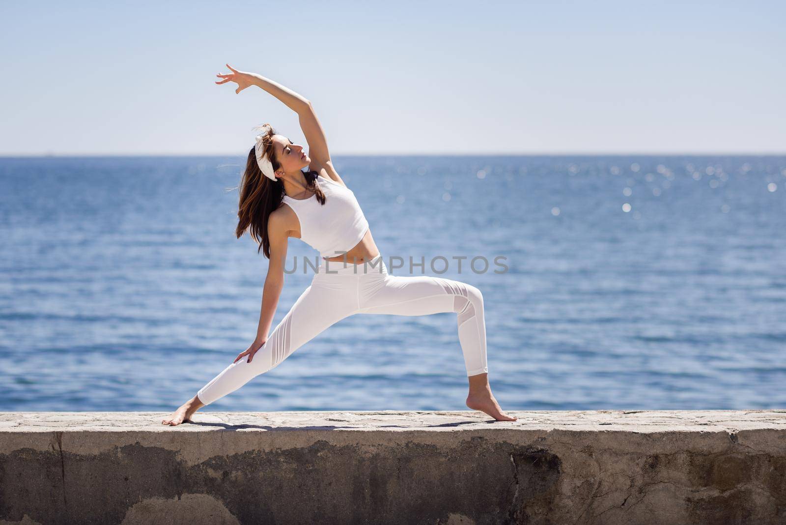 Young woman doing yoga in the beach. Female wearing white sport clothes in warrior at the sun figure with defocused sea background.