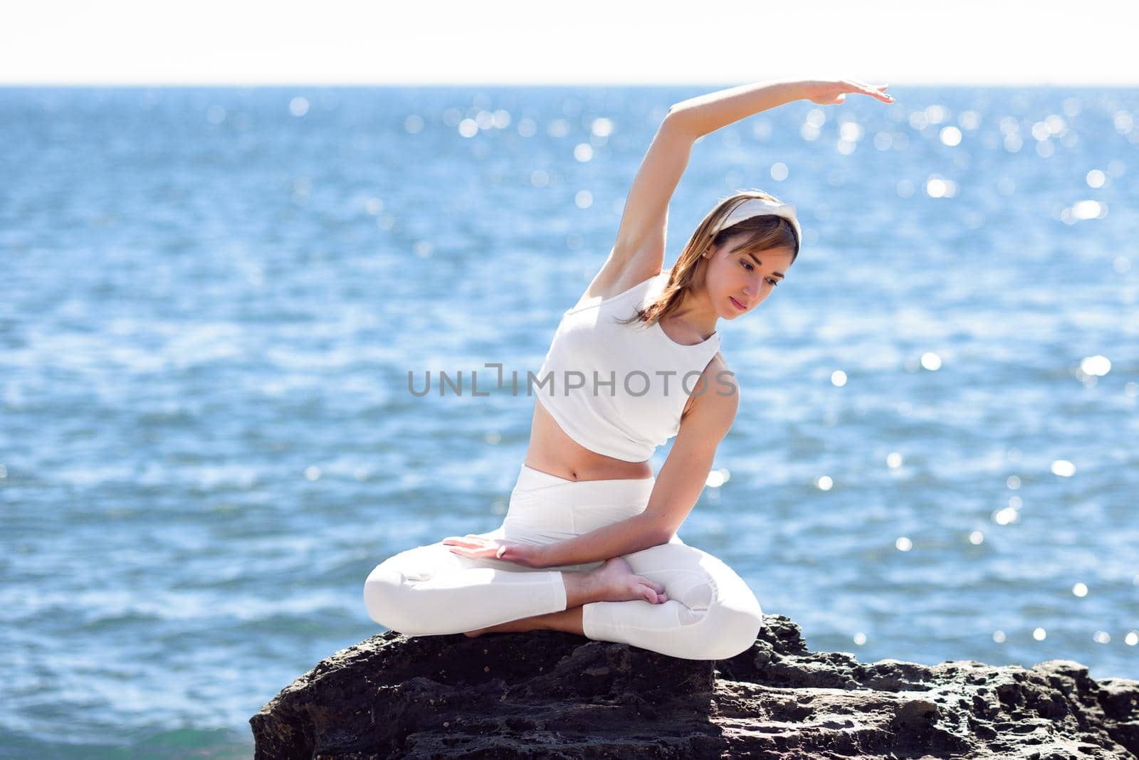 Young woman doing yoga in the beach. Female wearing white sport clothes in lotus figure with defocused sea background.