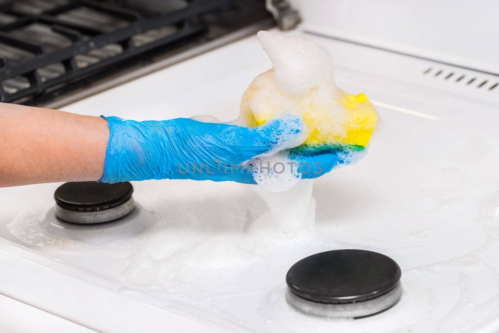 Woman's hand in a household glove washing sponge with foam a dirty gas stove background, close up by AYDO8