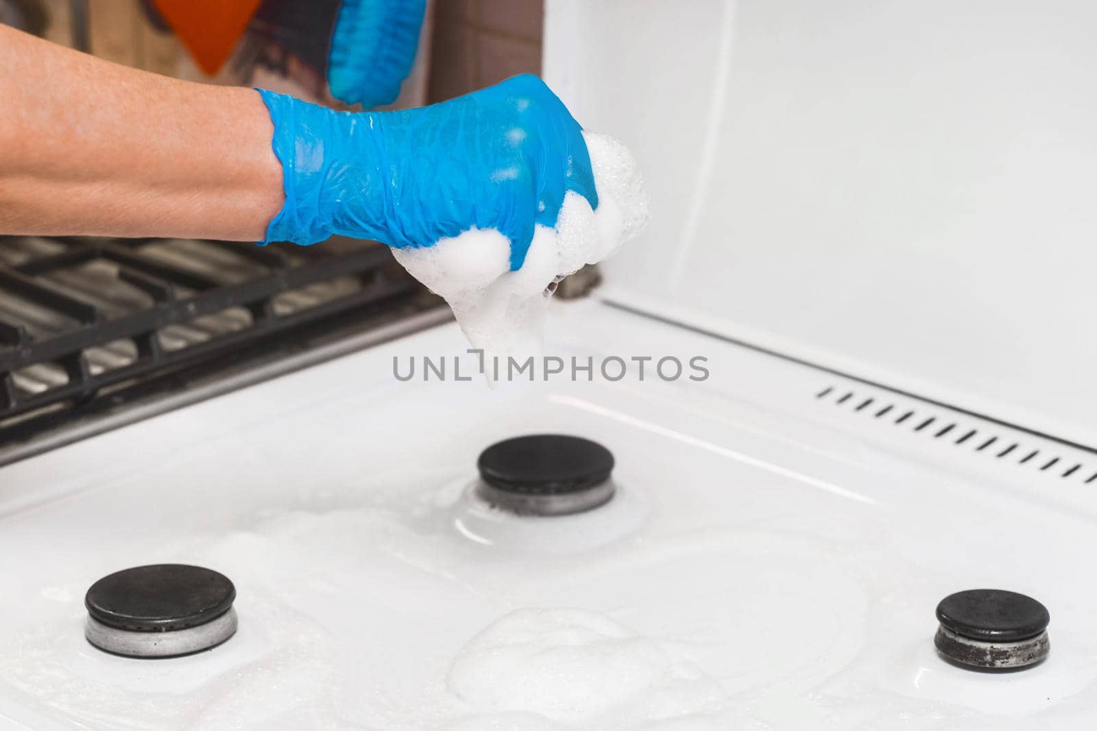 The hand of a housewife girl in a latex household glove squeezes a sponge with foam over a gas stove and burners in the kitchen, close-up by AYDO8