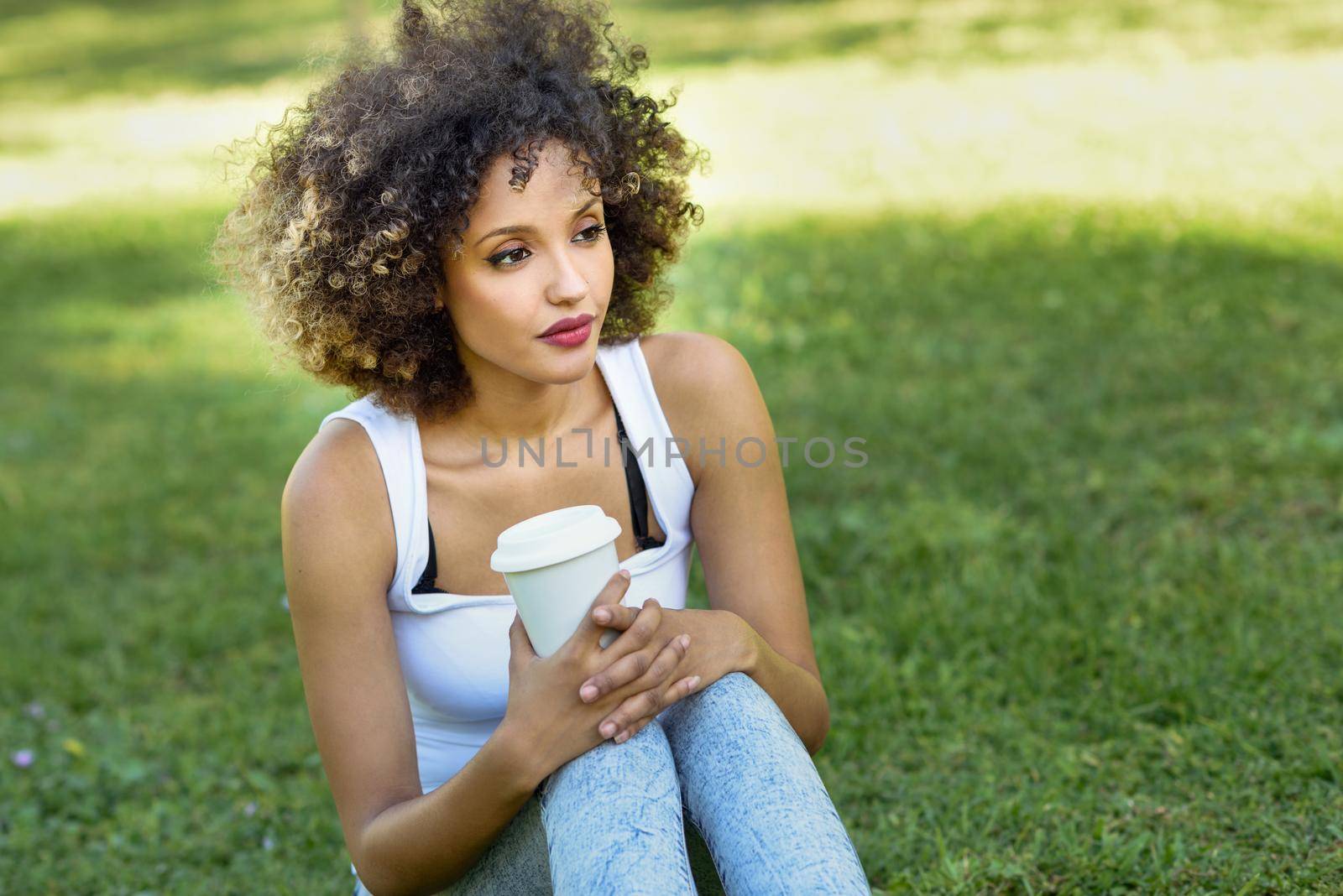 Beautiful young African American woman with afro hairstyle. Girl drinking coffee with a take away glass in park sitting on grass wearing casual clothes smiling.