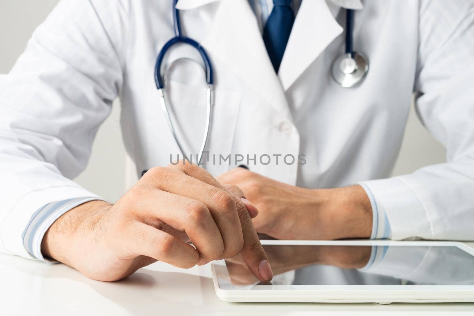 Close up of male doctor hands using tablet computer. Therapist in white medical gown with stethoscope sitting at desk. Modern technology in diagnosis and treatment. Medical digital application.