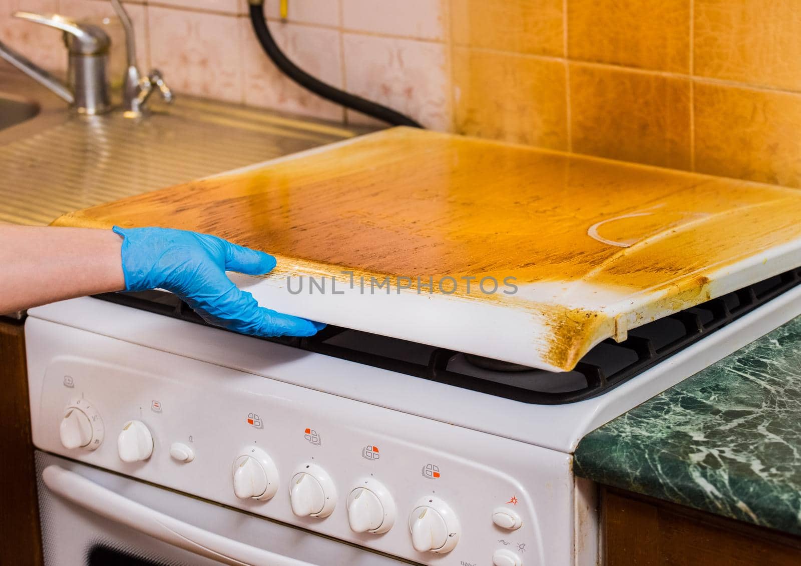 A woman's hand of a housewife in a household rubber glove holds the lid of a dirty gas stove with a sticky coating. Home cleaning of the kitchen.