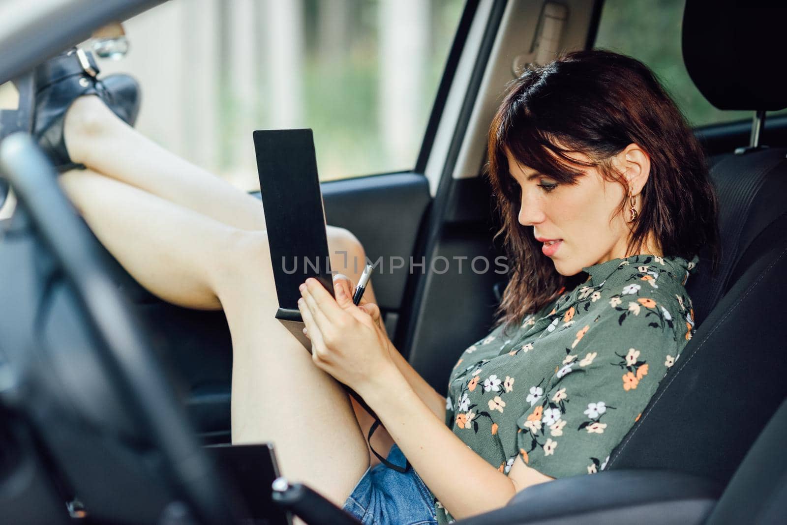 Young woman writing in a notebook with a pen in a white car pulling her feet out the window. Female wearing flowered shirt and shorts.