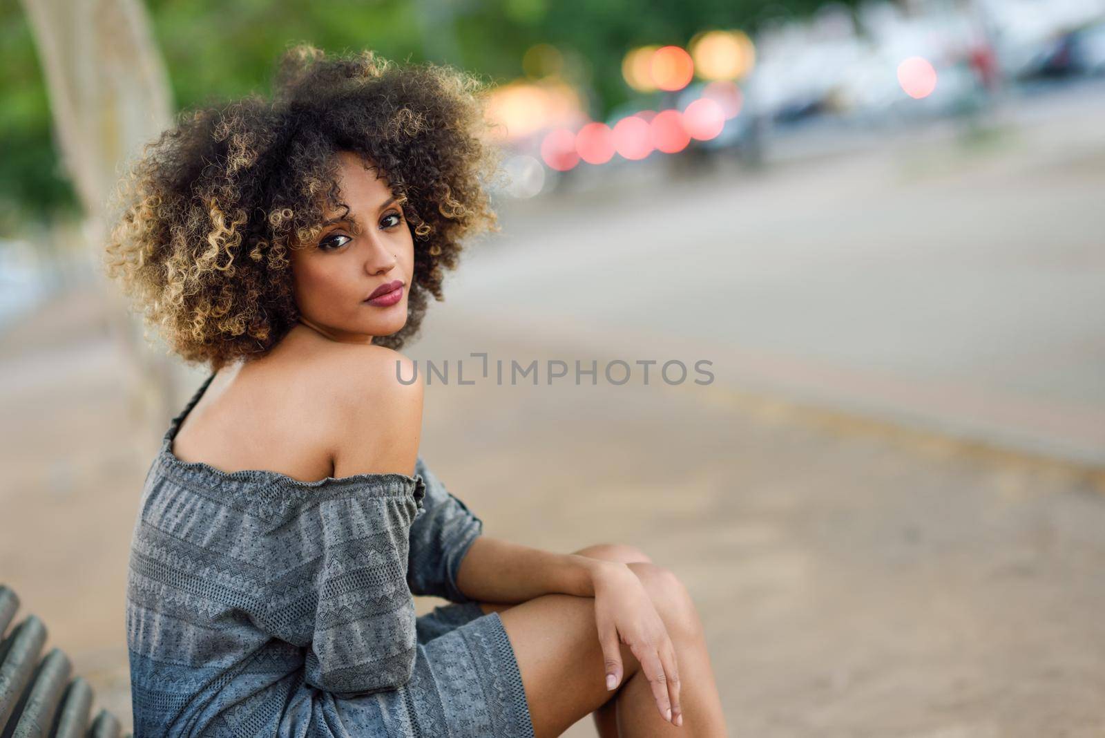 Young black girl with afro hairstyle sitting on a bench in urban background. Mixed woman wearing casual clothes