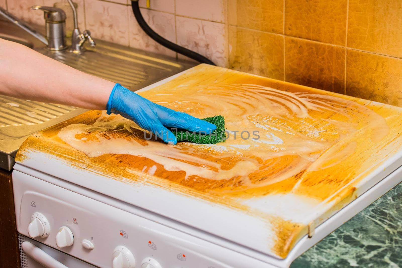 Woman's housewife hand in a household glove washes a dirty coating on a gas stove on kitchen background, close up.