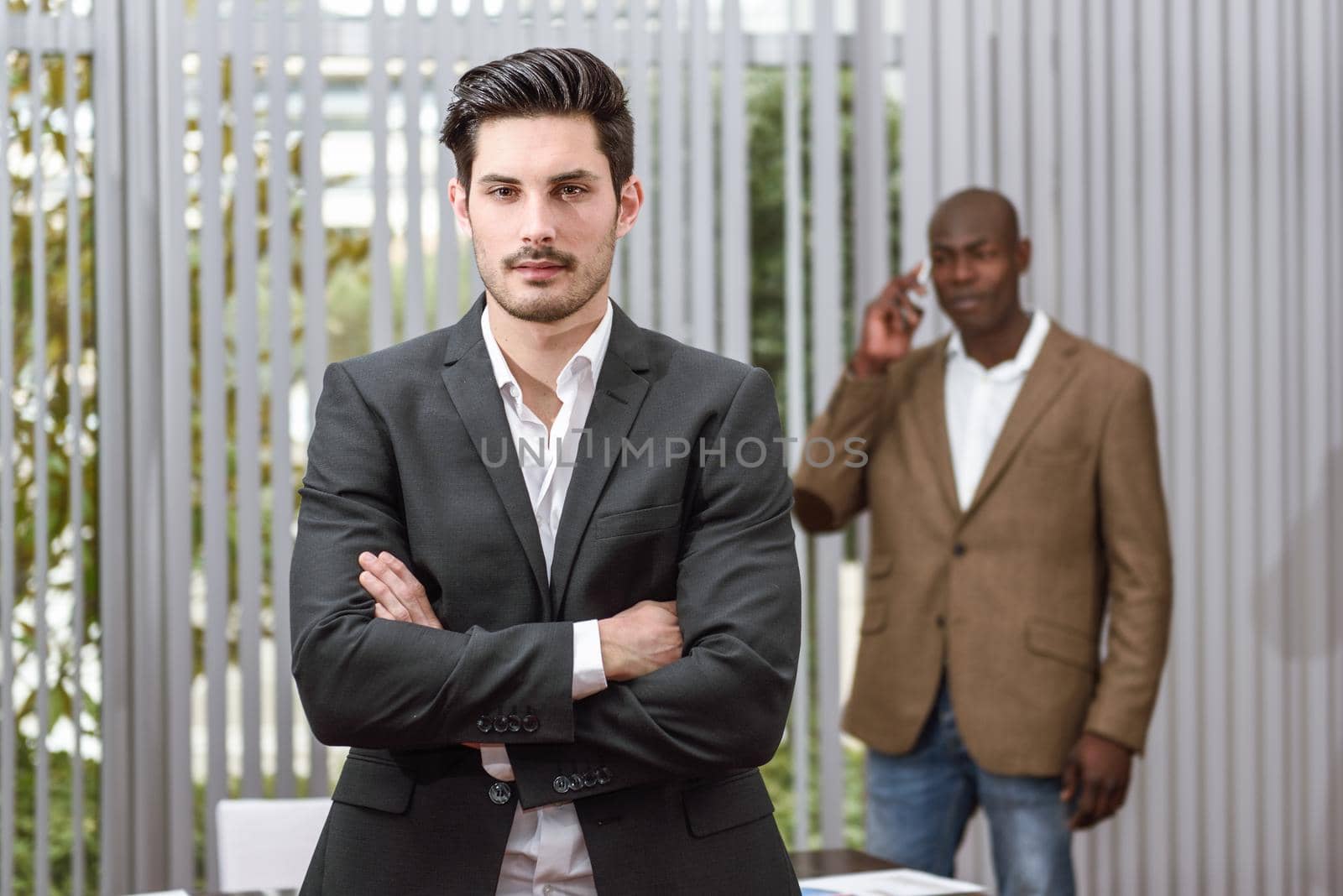 Confident businessman. Cheerful young caucasian man in formalwear keeping arms crossed and smiling while his black colleague standing on background using a smart phone