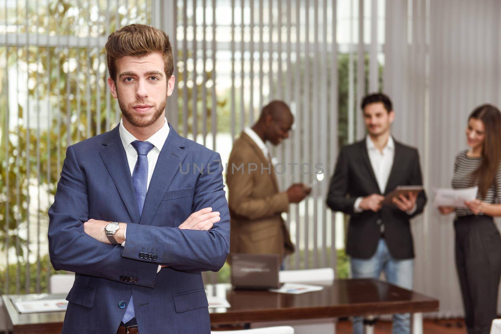 Businessman leader looking at camera with arms crossed in working environment. Group of people in the background