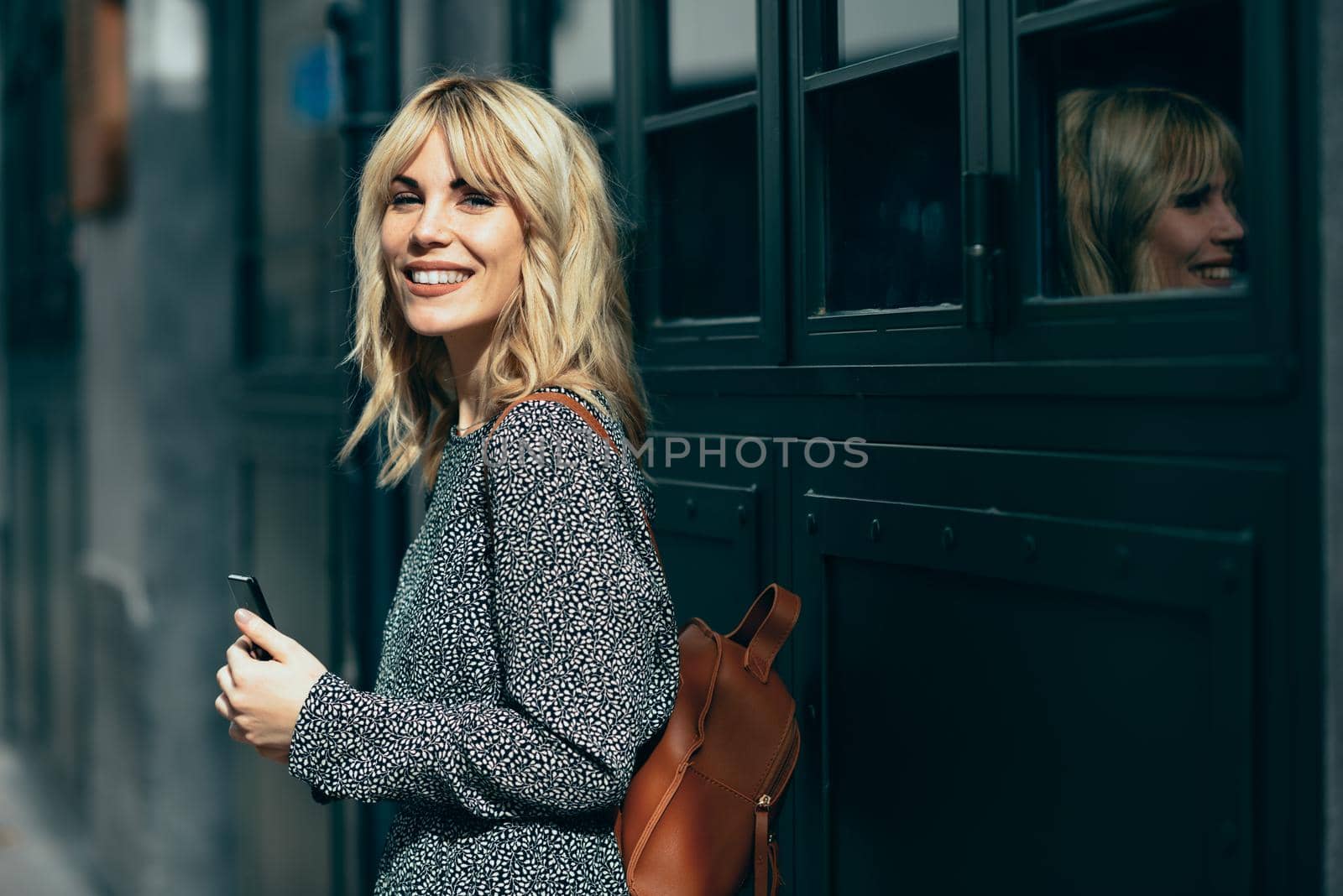 Attractive young caucasian woman looking at her smart phone in urban background. Blonde girl wearing casual clothes outdoors