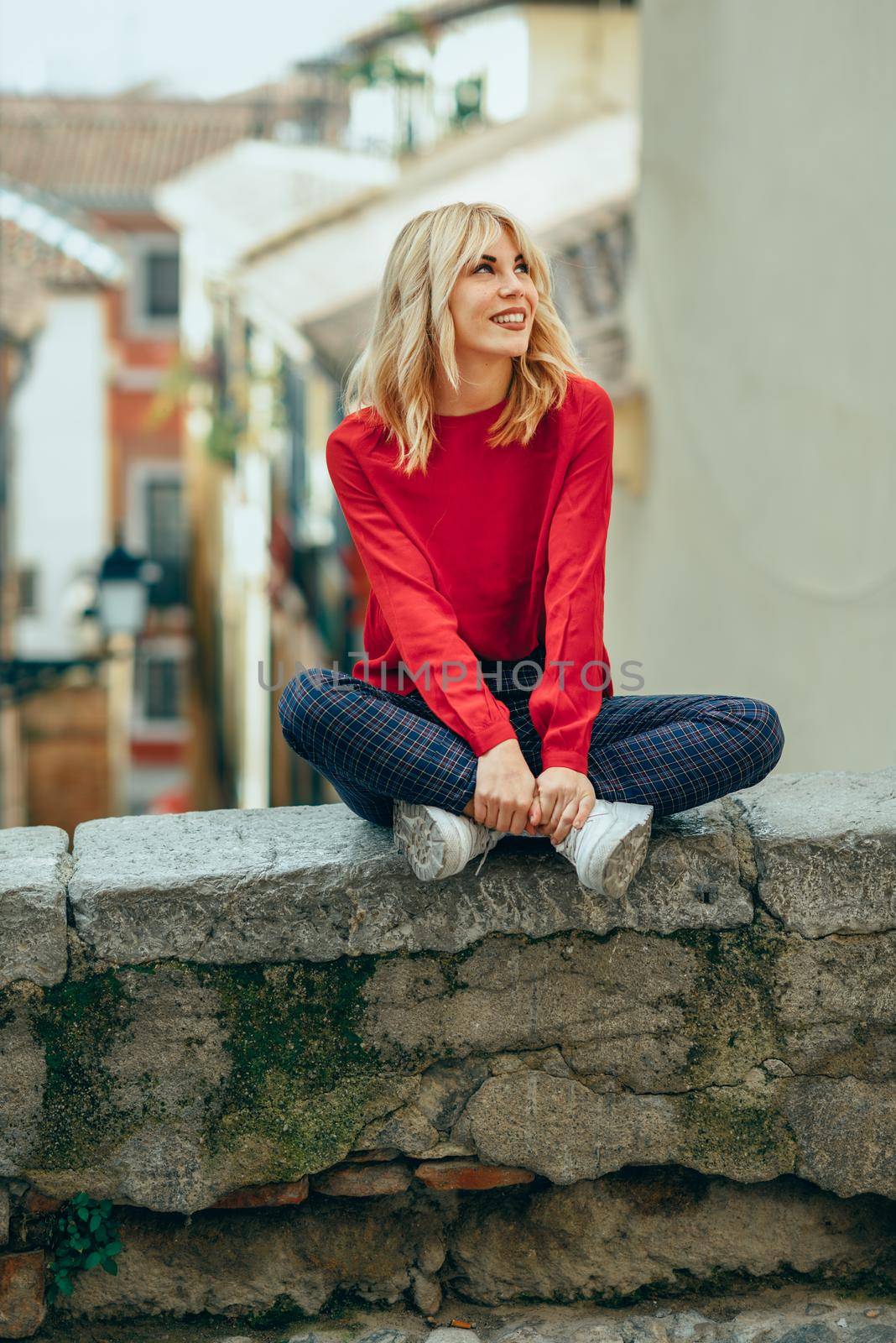 Happy young blond woman sitting on urban background. Smiling blonde girl with red shirt enjoying life outdoors.