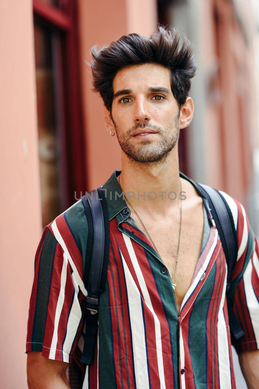 Attractive young man with dark hair and modern hairstyle wearing casual clothes in urban background.