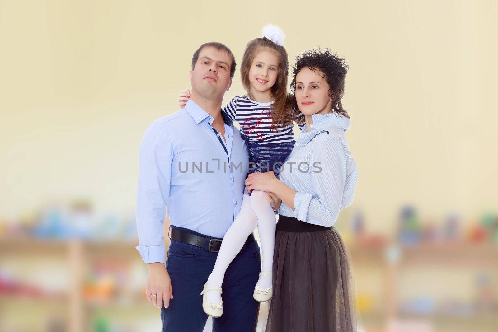 Happy young family, mom dad and little daughter.Parents keep the girl in her arms , and she hugs their neck.In the Montessori room the children's garden where there are shelves with toys and material.