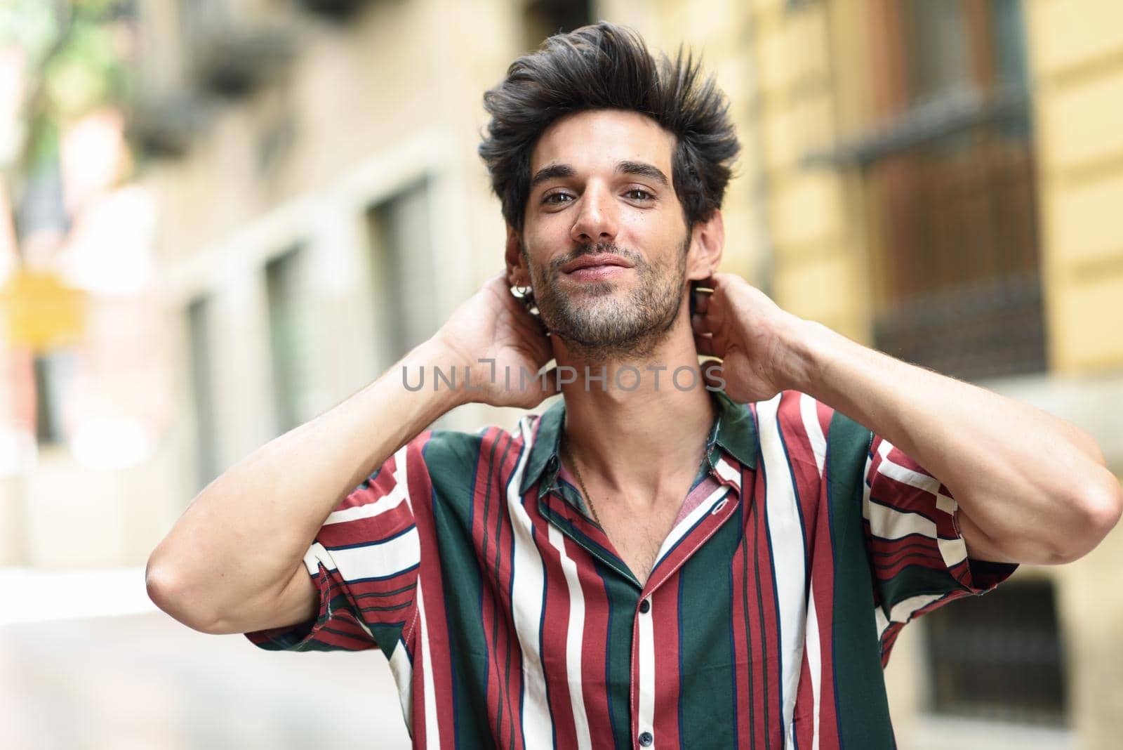 Attractive young man with dark hair and modern hairstyle wearing casual clothes in urban background.