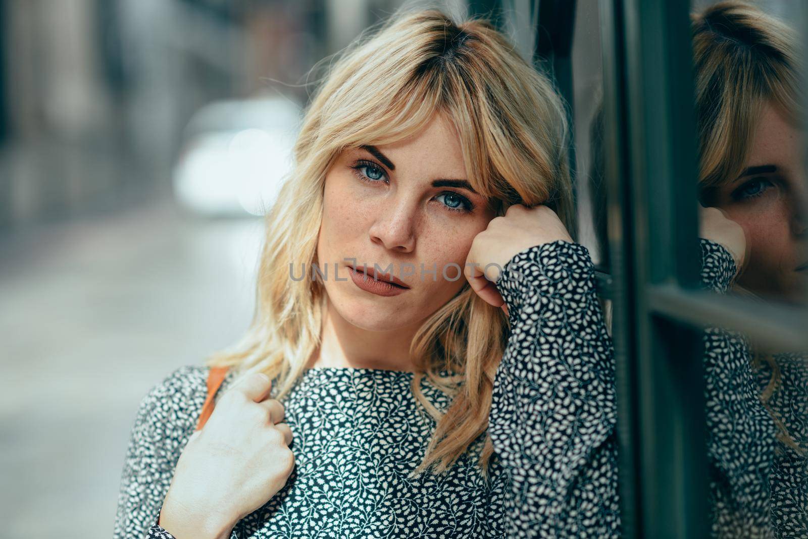 Portrait of serious young blonde woman standing on urban background. Beautiful girl with blue eyes wearing dress standing outdoors.