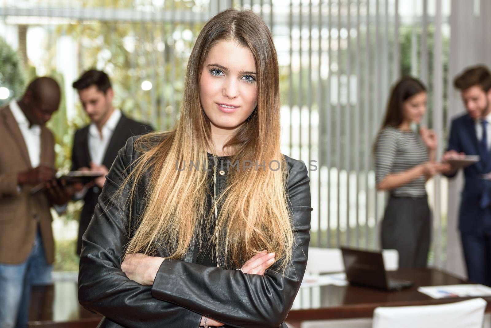 Blonde businesswoman leader looking at camera with arms crossed in working environment. Group of people in the background