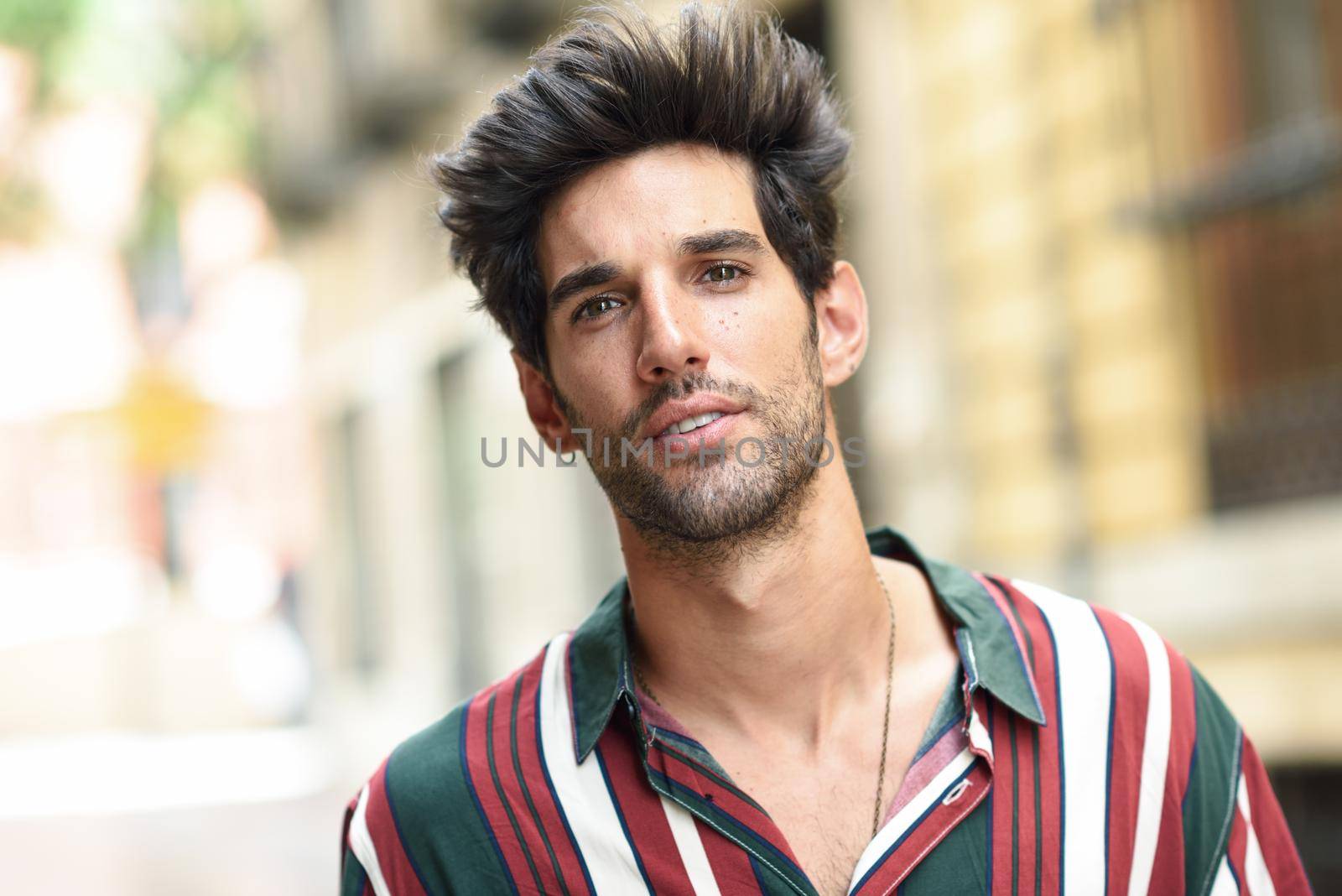 Attractive young man with dark hair and modern hairstyle wearing casual clothes in urban background.