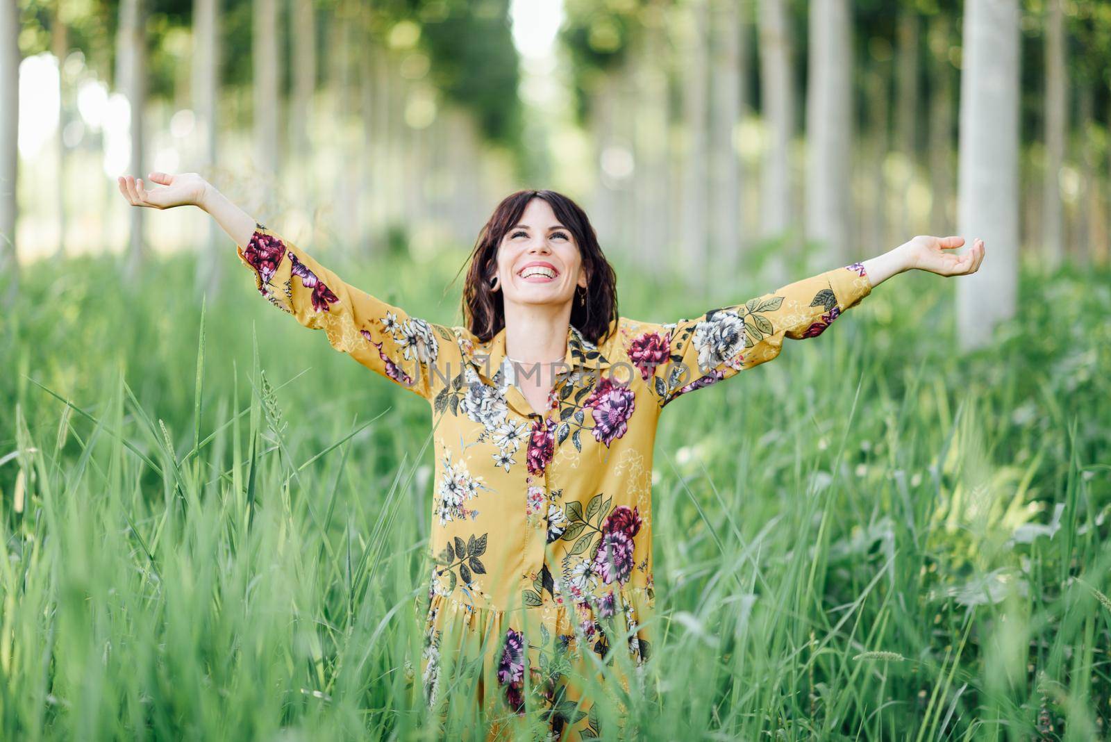 Enjoying the nature. Young woman arms raised enjoying the fresh air in green forest. Female wearing flowered dress.