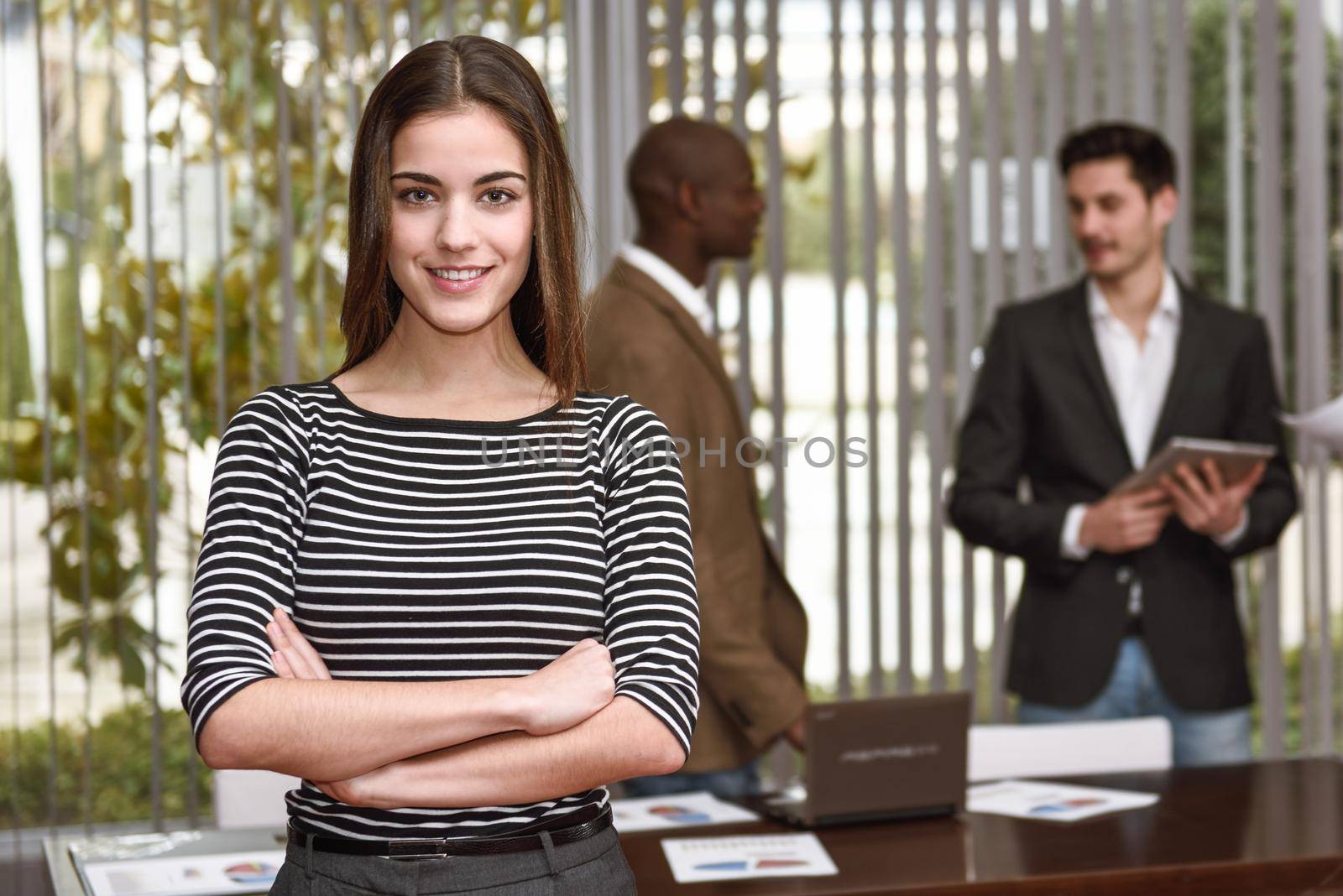 Young businesswoman leader looking at camera with arms crossed in working environment. Group of multiracial people in the background