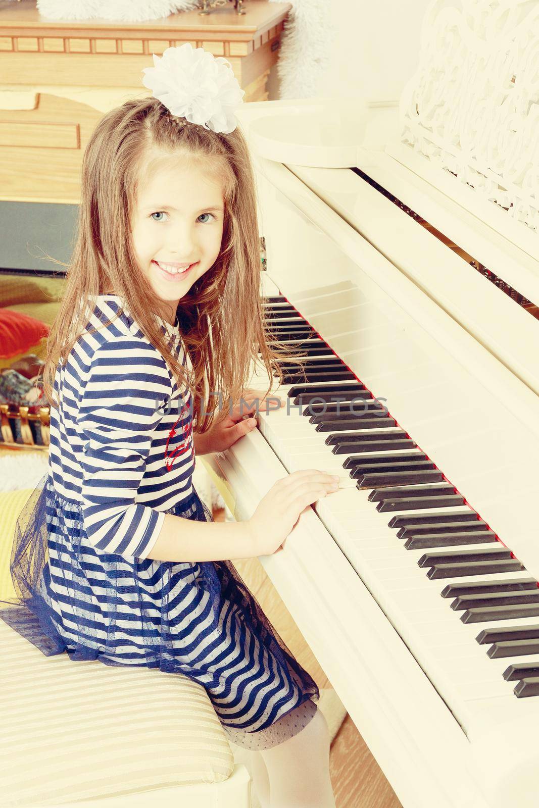 Little girl in blue striped dress and a white bow on her head.Girl smiling sitting behind the keys of a large white piano.Creative toning of a photograph.