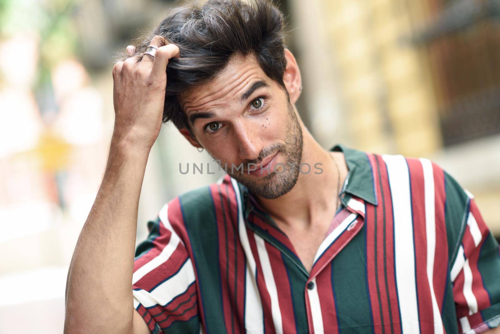 Attractive young man with dark hair and modern hairstyle wearing casual clothes in urban background.