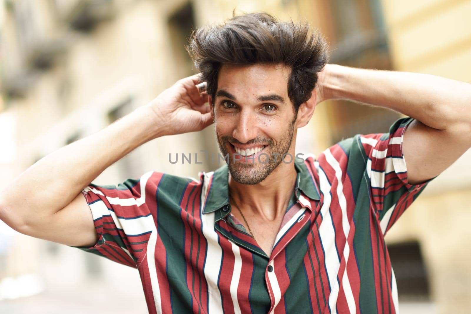 Smiling young man with dark hair and modern hairstyle wearing casual clothes in urban background.