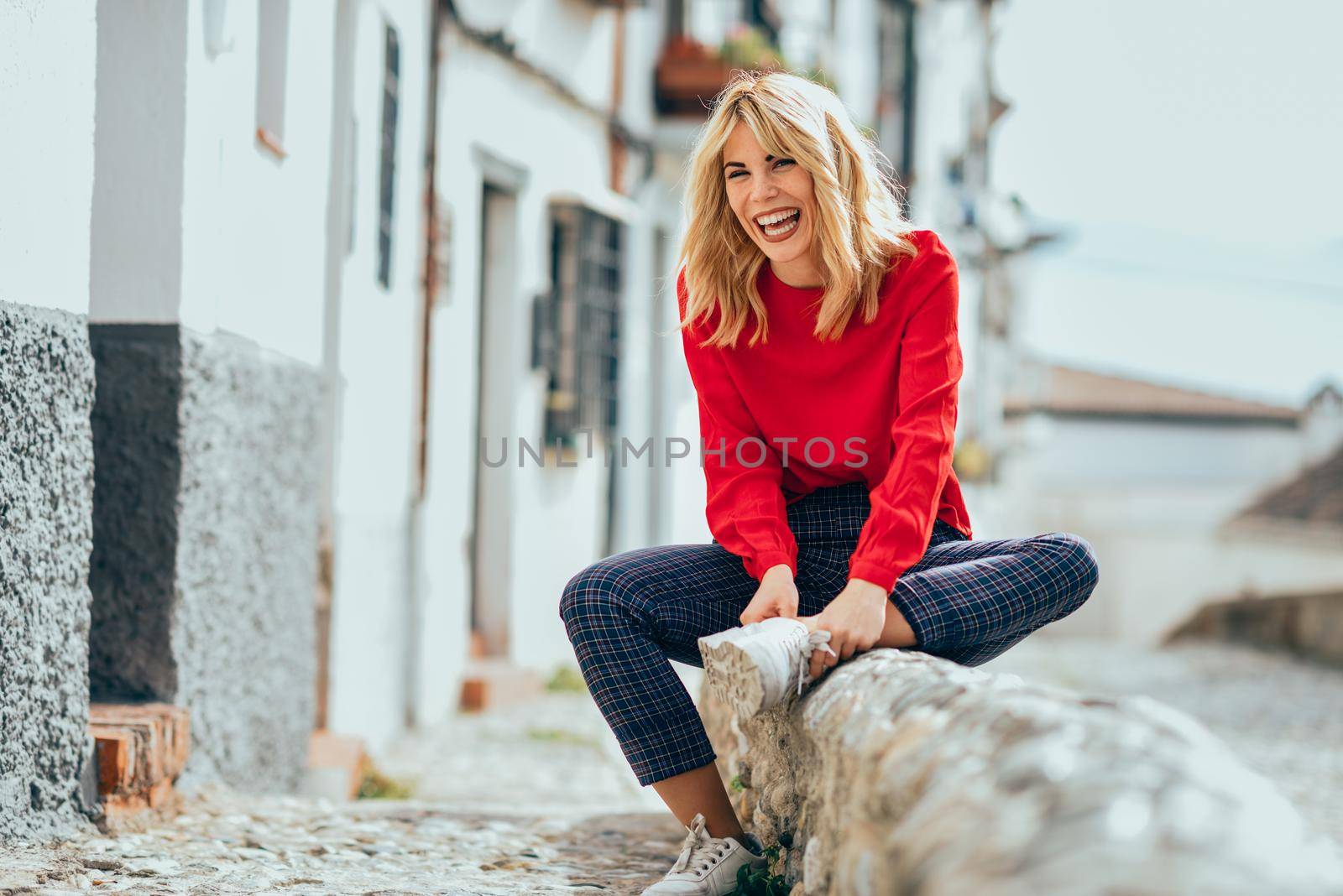 Happy young blond woman sitting on urban background. Laughing blonde girl with red shirt enjoying life outdoors.