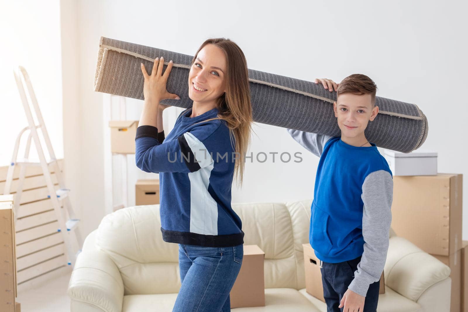 Smiling caucasian mother and a charming son are holding a folded carpet on their shoulders in a new living room intending to spread it in a new apartment by Satura86