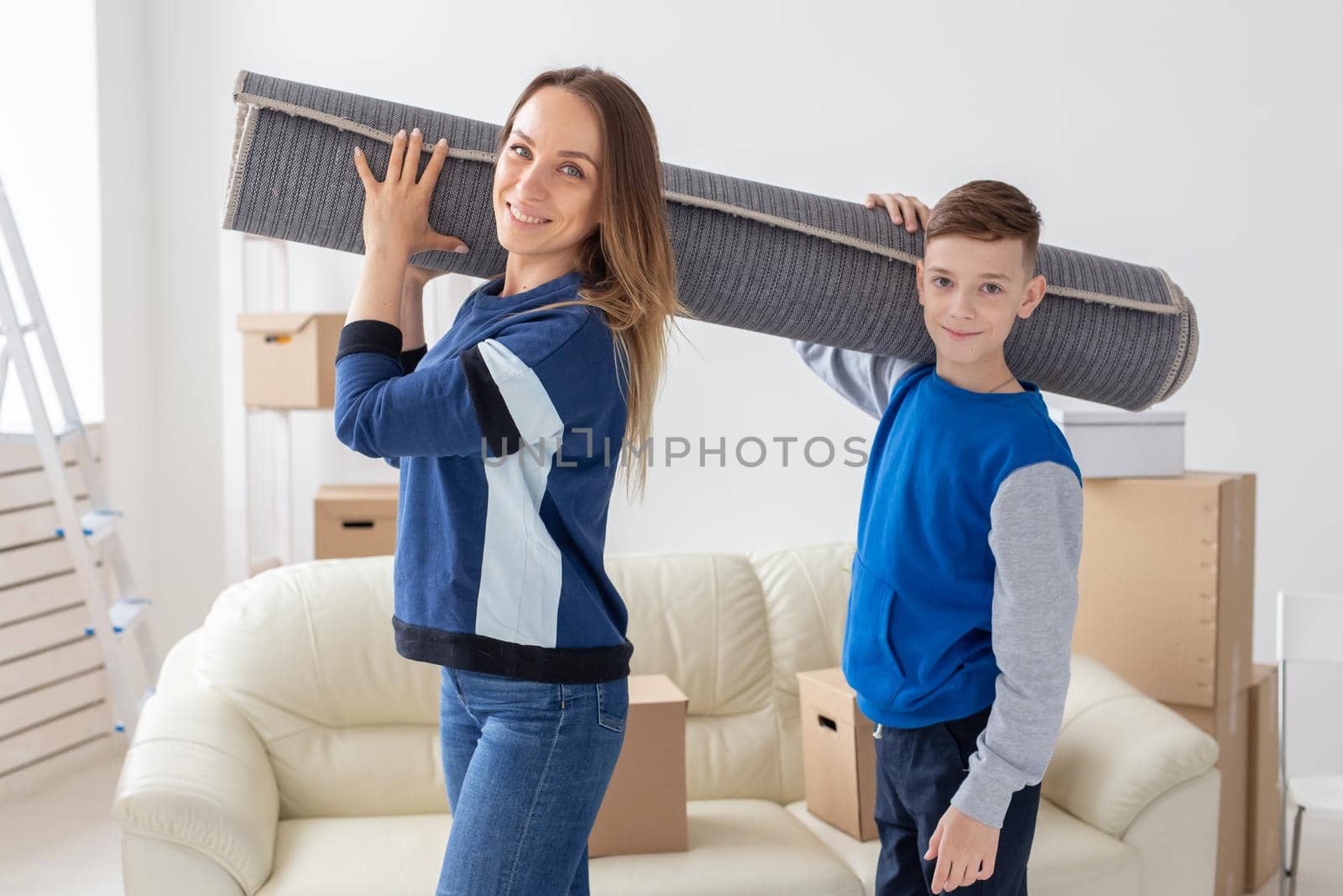 Smiling caucasian mother and a charming son are holding a folded carpet on their shoulders in a new living room intending to spread it in a new apartment. Housewarming concept