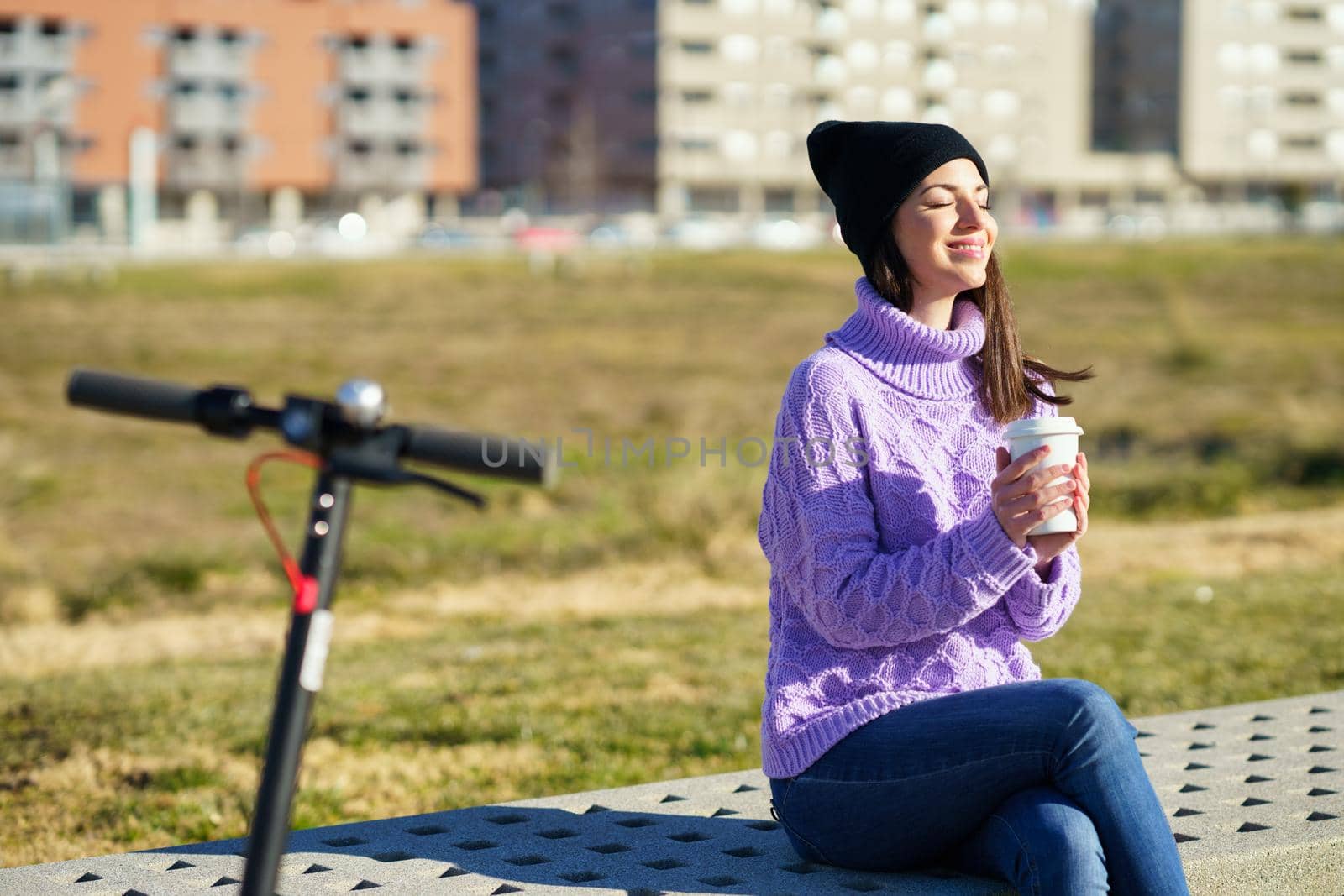 Female student with electric scooter taking a coffee break enjoying the winter sunlight with her eyes closed. Lifestyle concept.