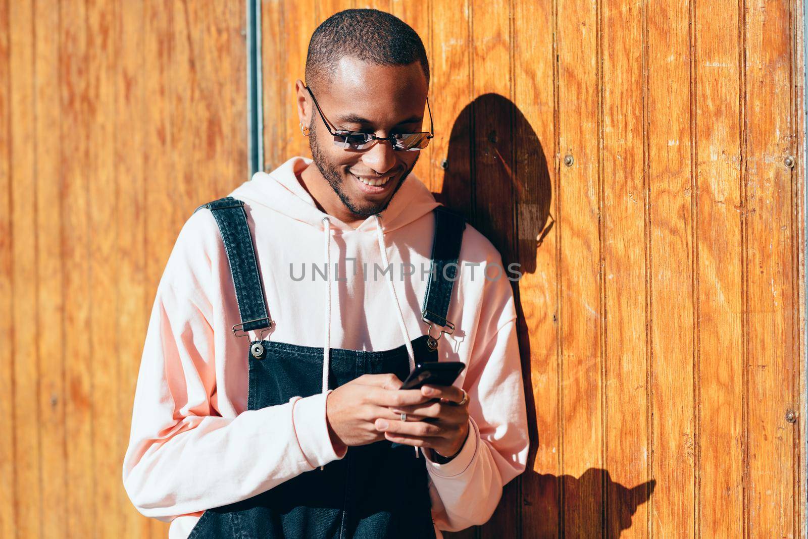 Young black man wearing casual clothes and sunglasses using smart phone against a wooden background. Millennial african guy with bib pants outdoors smiling