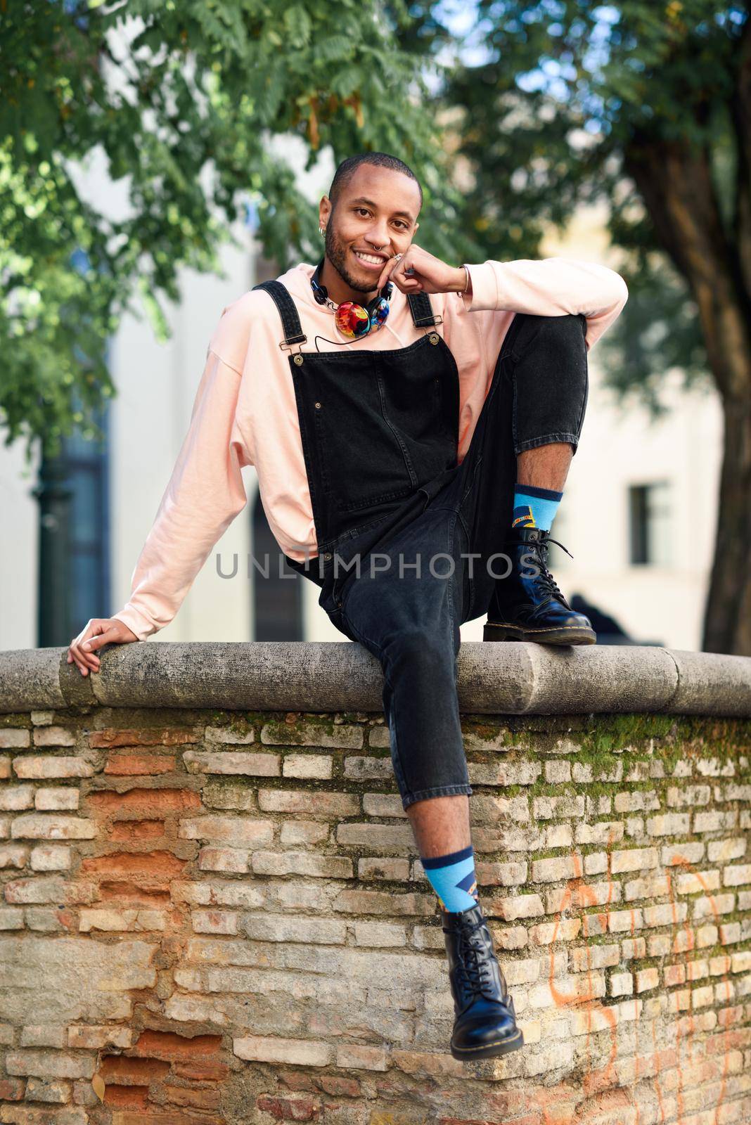 Young black man wearing casual clothes and headphones sitting on urban background. Happy African guy with bib pants outdoors