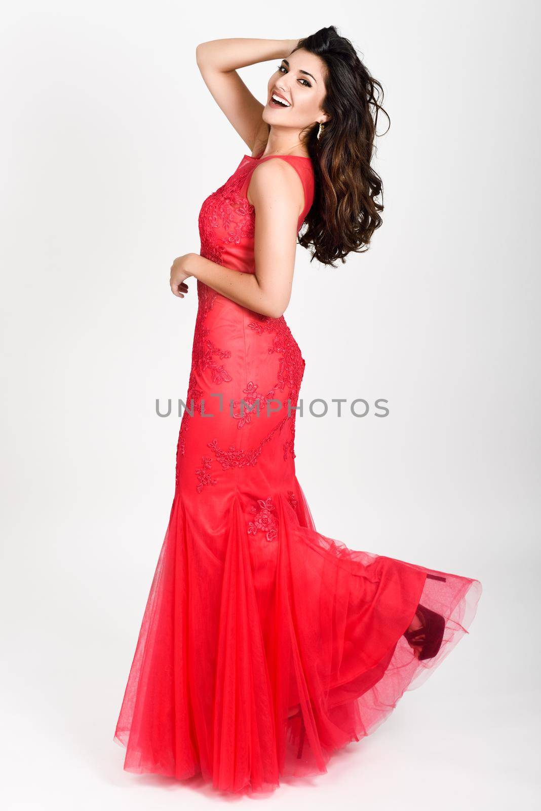 Young woman wearing long red dress on white background. Brunette girl with long hair and wavy hairstyle smiling to camera.