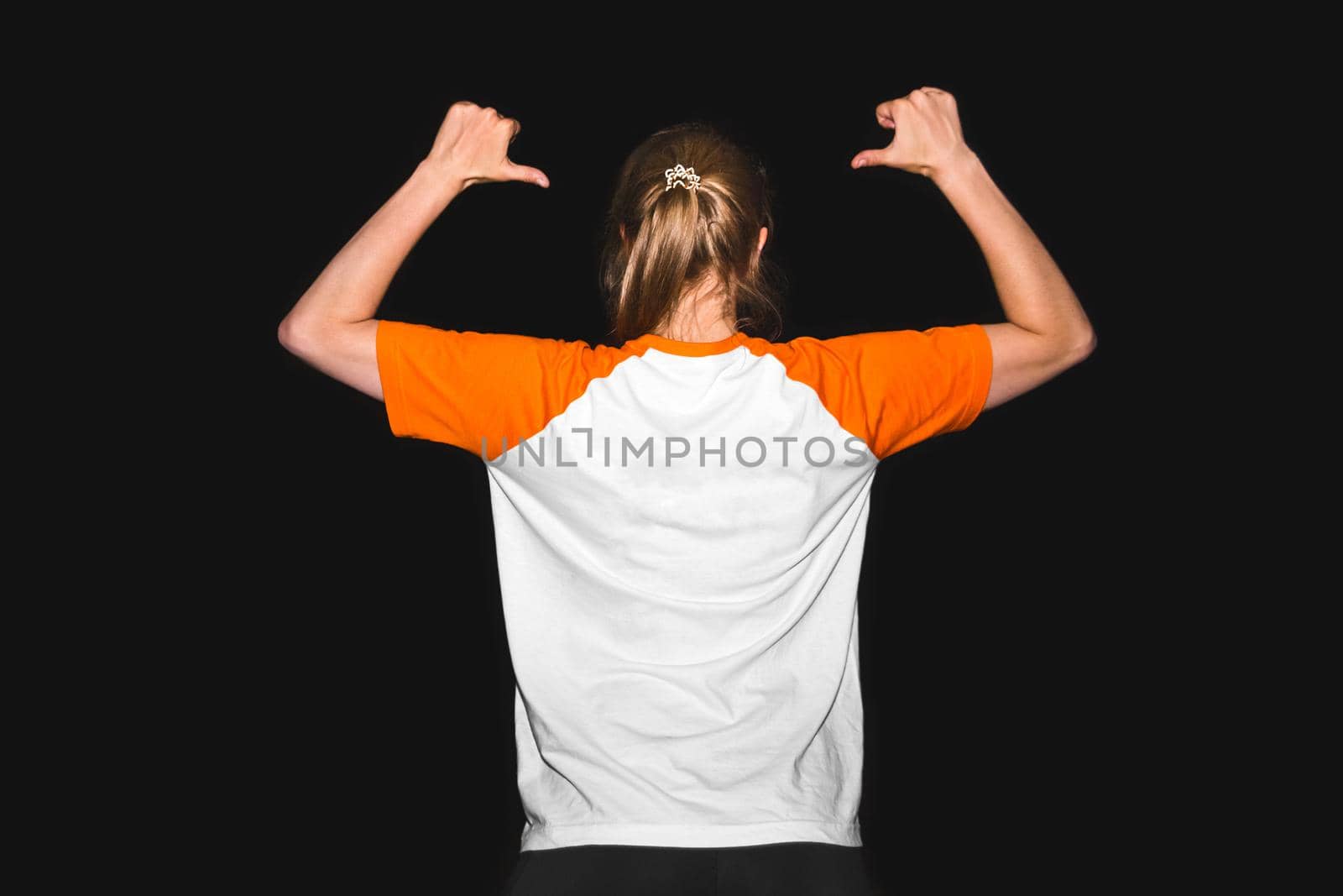 A girl in a T-shirt on a black background points with her hands to her back, a place for text. Design copy space clothes by AYDO8