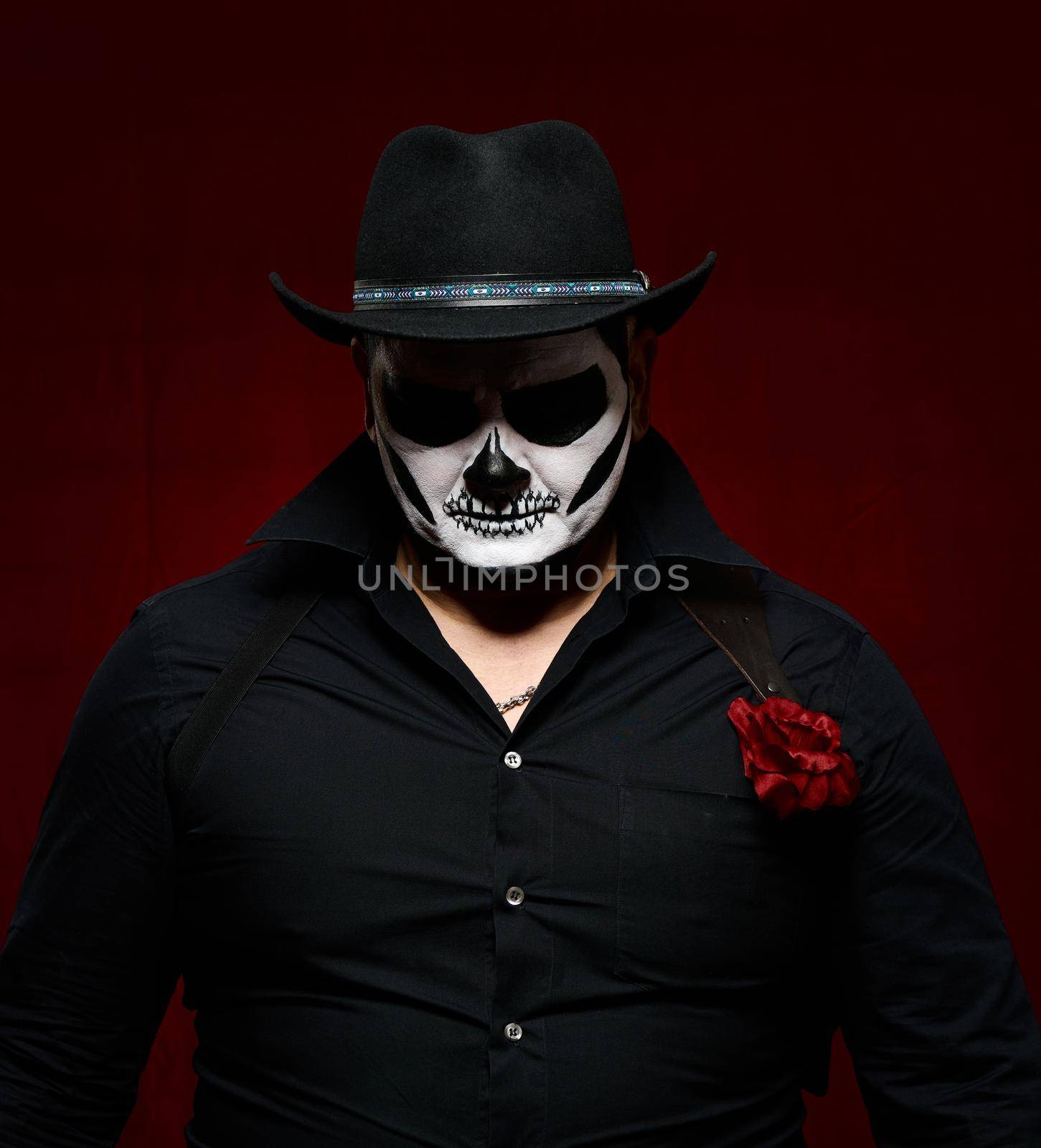 man is painted with a skeleton in a black hat and shirt, head down, red background