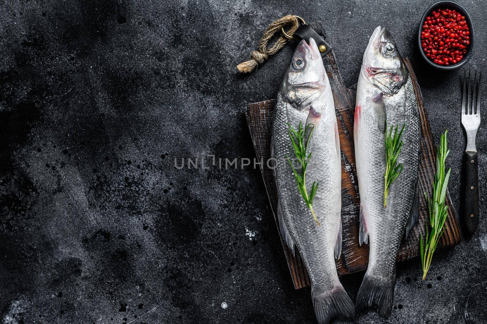 Raw seabass fish on a cutting board with spices, herbs. Black background. Top view. Copy space.