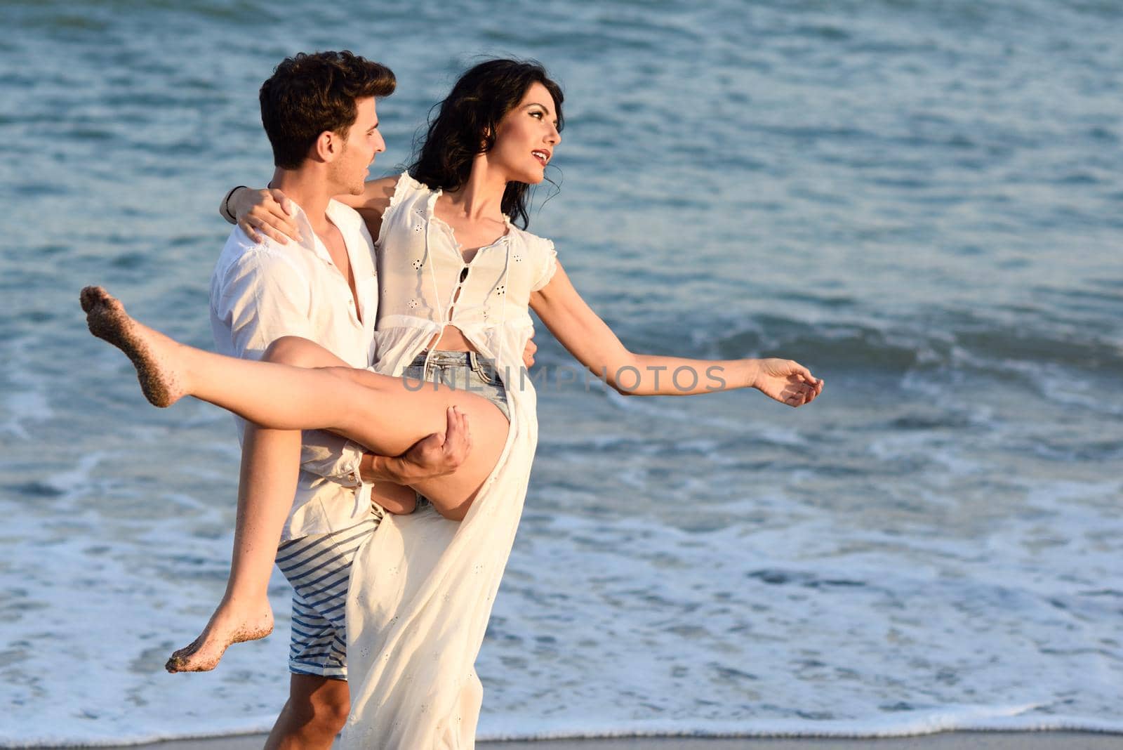 Young happy couple walking in a beautiful beach. Funny Man and woman wearing casual clothes. Male carrying his girlfriend in his arms.