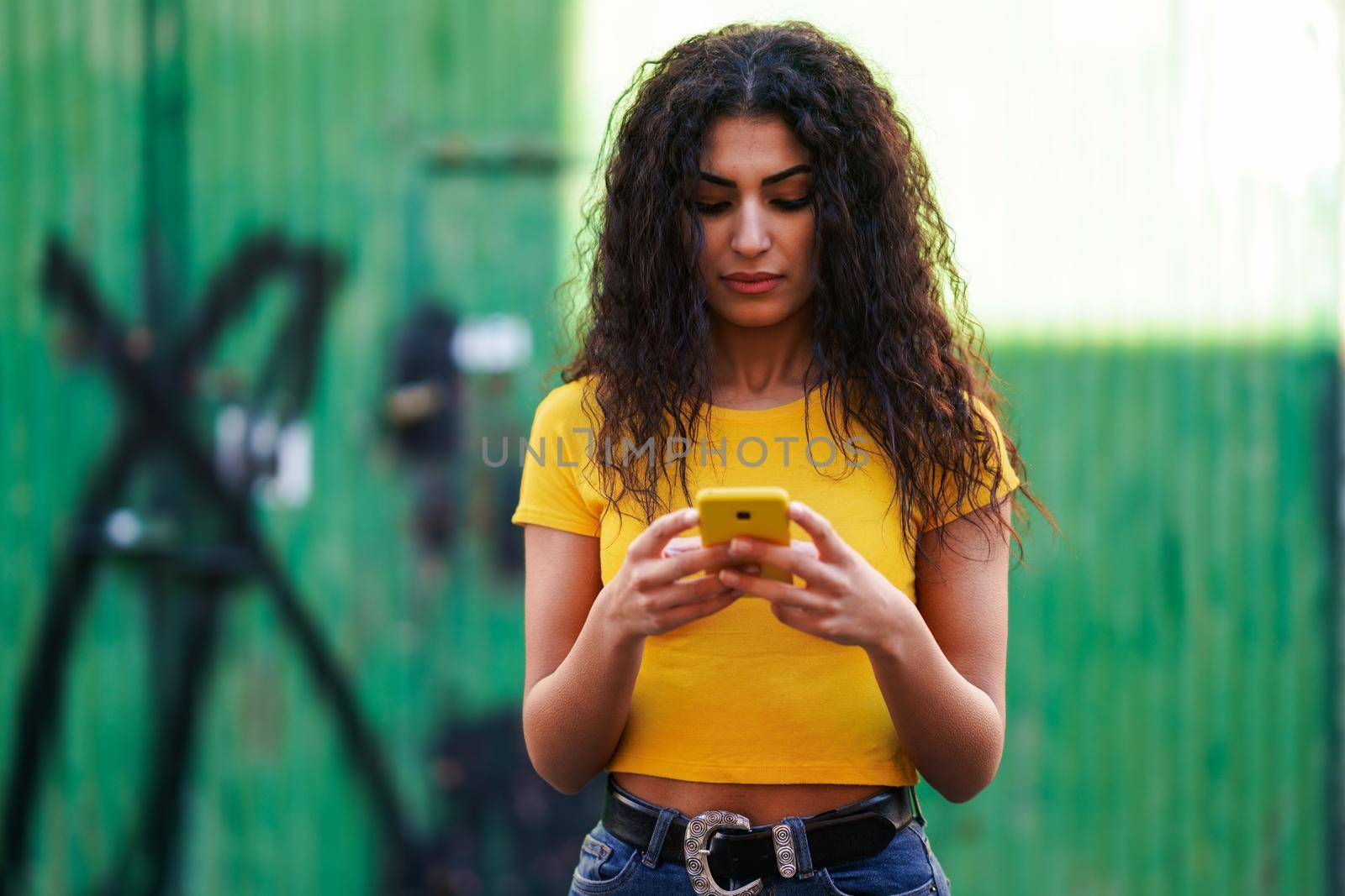 Young Arab woman texting a text message with her smartphone against green urban wall.