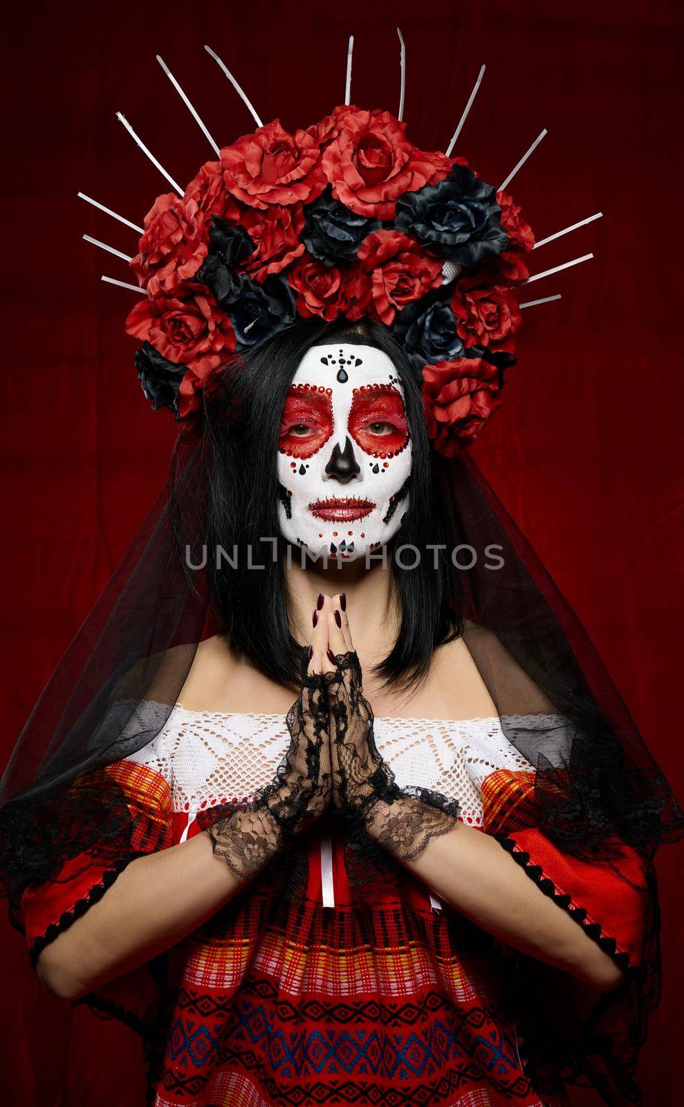 beautiful woman with a sugar skull makeup with a wreath of flowers on her head and a skull hands raised up in prayer pose in black gloves, red background