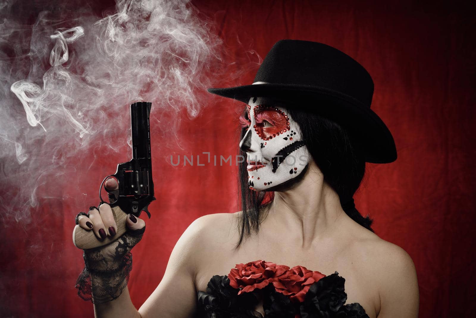 beautiful woman in skeleton make-up holds a revolver, she is wearing a black hat. White smoke over red background from shot by ndanko