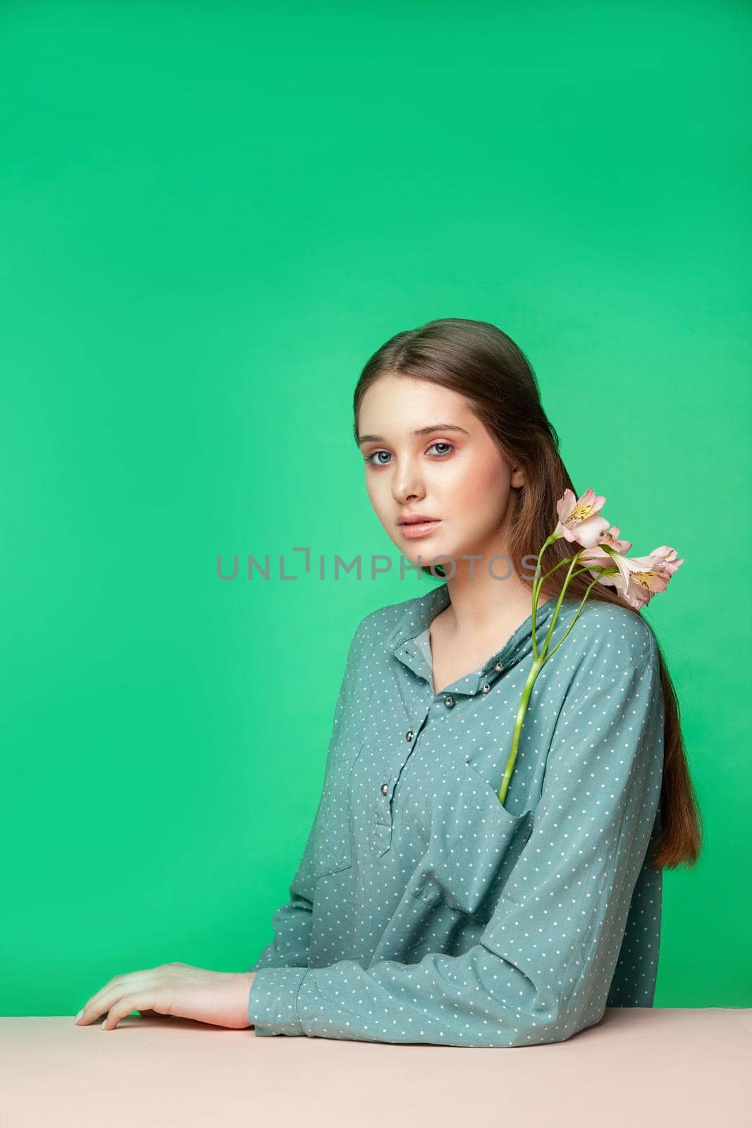 Young woman with fresh flowers in pocket of stylish dotted shirt looking at camera while sitting at table against green background