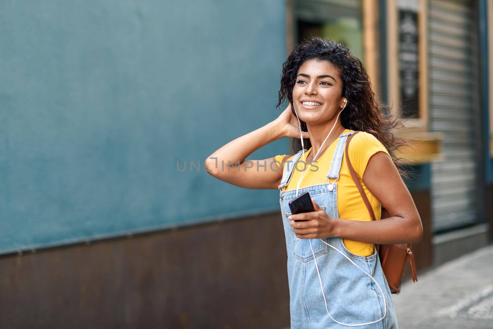 Attractive African woman listening to music with earphones outdoors. Black girl in casual clothes with curly hairstyle in urban background.