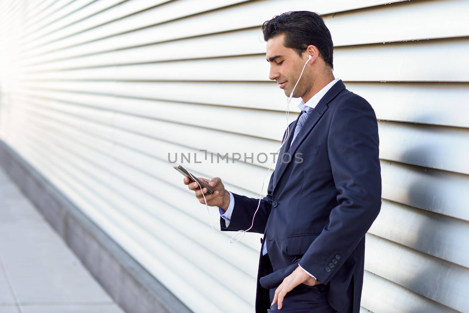 Young businessman wearing blue suit and tie using a smartphone in urban background. Man with formal clothes and headphones in the street.