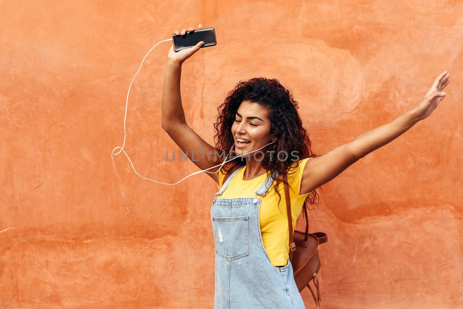 Happy Arab girl listening to music and dancing with earphones outdoors. African woman in casual clothes with curly hairstyle in urban background.