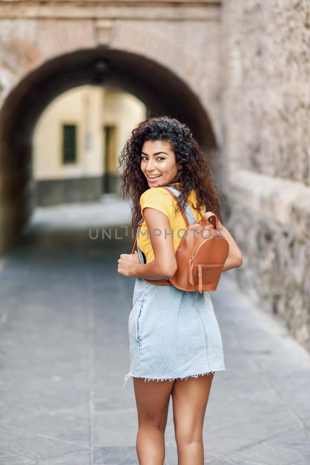 Rear view of young Arab woman with backpack outdoors. Traveler girl in casual clothes in the street. Happy female wearing yellow t-shirt and denim dress in urban background.