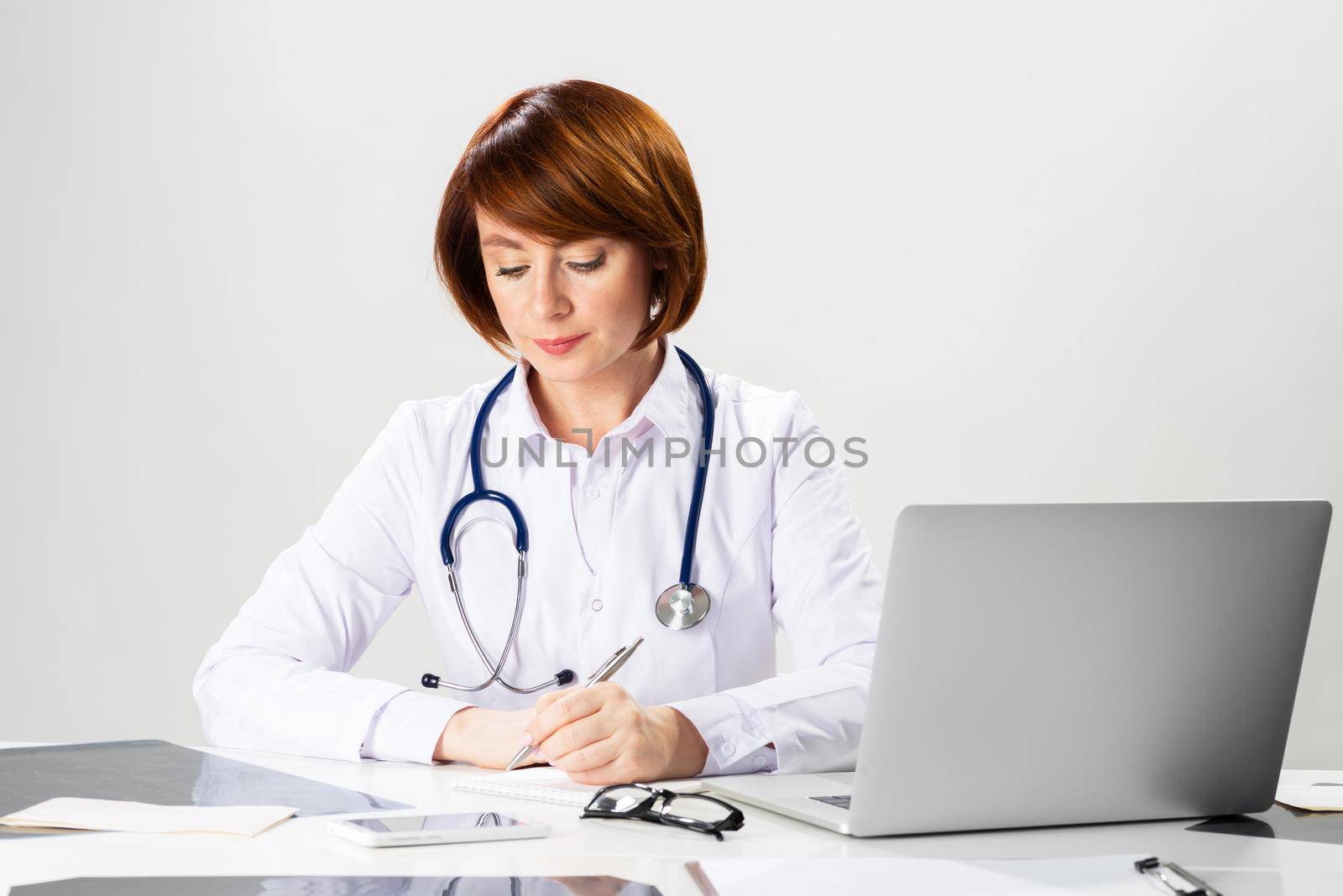 Beautiful redhead doctor writing on paper in office. Physician in white coat with stethoscope sitting at desk. Professional medical diagnosis and treatment in clinic. Healthcare and medicine.